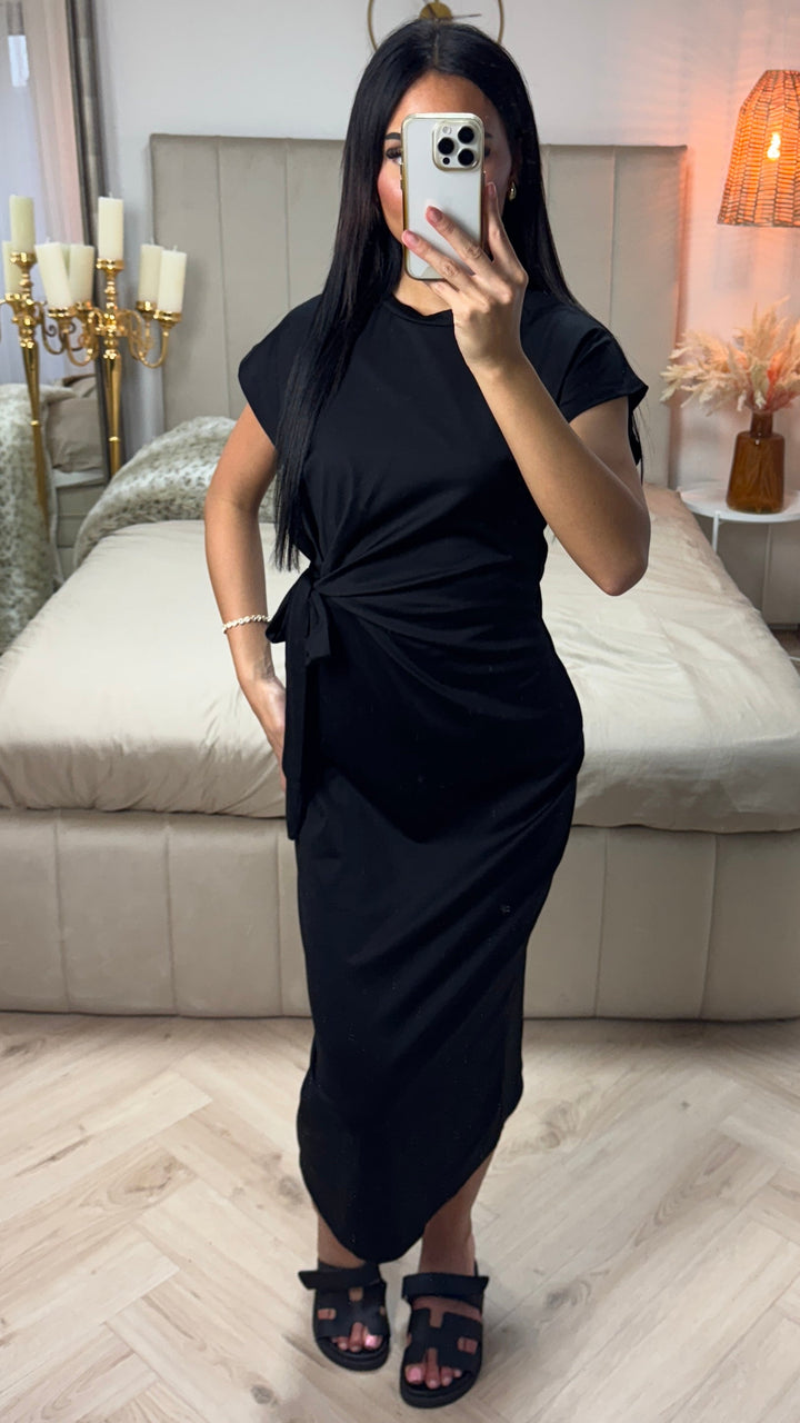 Front view of a model wearing the black Aurora midi dress with side bow detail and black sandals