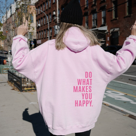 Back view, model in a pink relaxed knit pullover hoodie with "DO WHAT MAKES YOU HAPPY" text, street scene