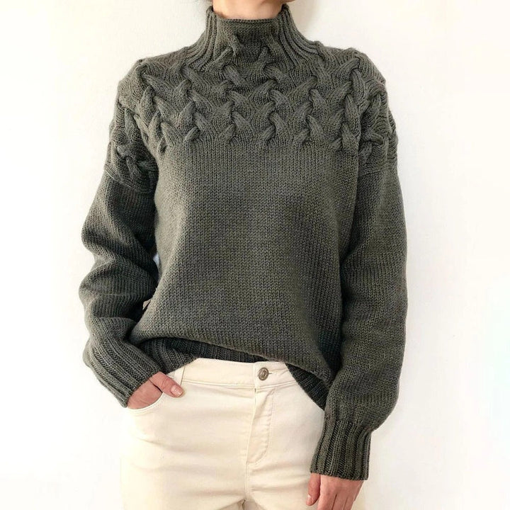 Front view of a model wearing a dark green cable knit mock neck sweater with cream trousers, hand in pocket