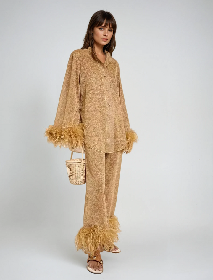 Front view, model wearing a gold Leona two-piece set with feather cuffs and hems, holding a small wicker bag