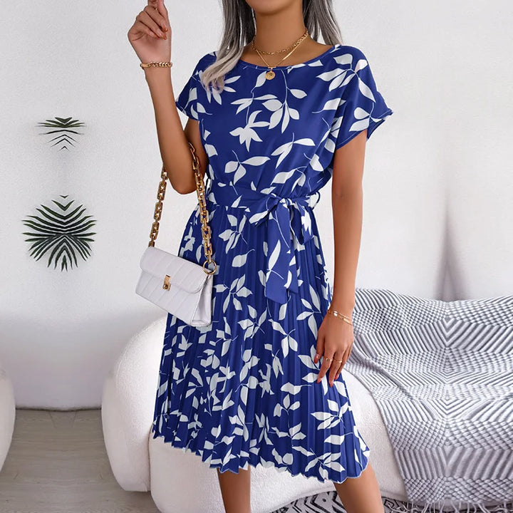 Front view of a model wearing a blue floral pleated maxi dress with white leaf print and a tie waist