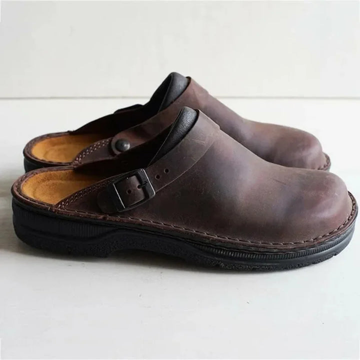 Three quarter view of a pair of brown PU leather slip-on shoes with black soles and buckle detail
