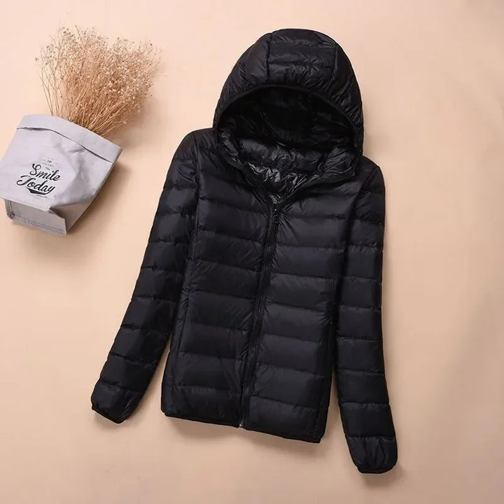 Front view of a black hooded lightweight puffer jacket with a zipper, next to dried flowers in a bag