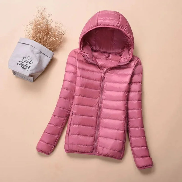 Front view of a pink hooded lightweight puffer jacket with a zipper, next to dried flowers in a bag