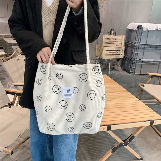 Smiley Print Corduroy Tote Bag worn by model, held over her shoulder, standing near a wooden table