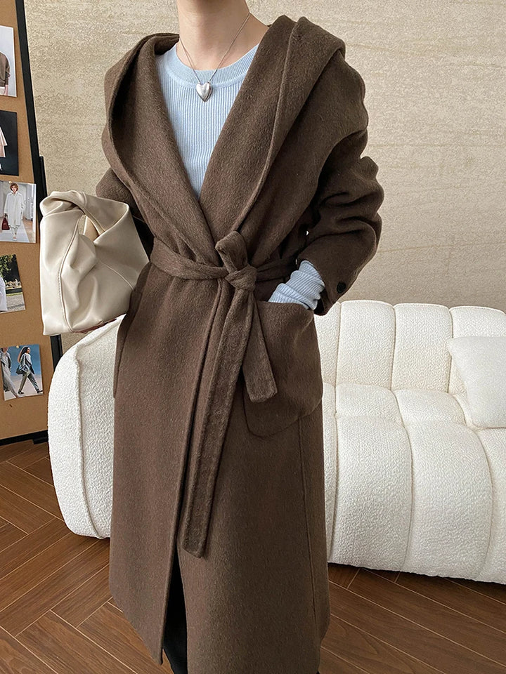 Front view, model wearing a long belted hooded wool blend wrap coat in brown with a cream bag