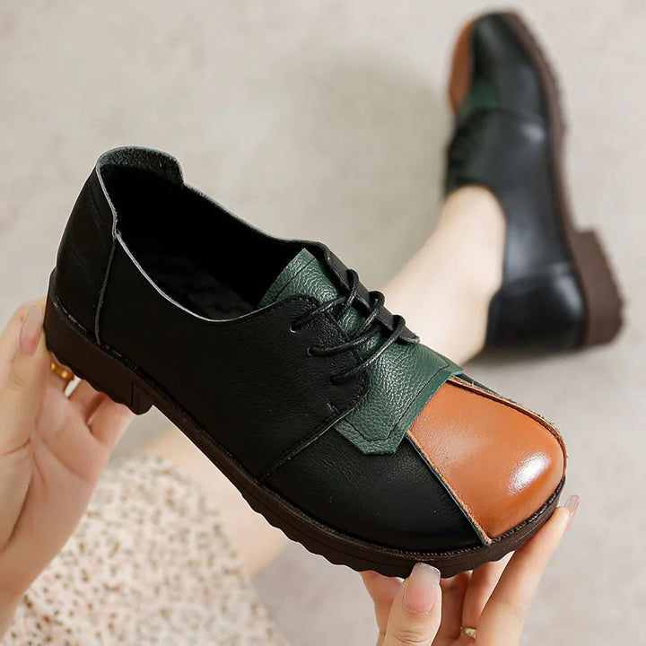 Side view of a Nora genuine leather flat, held by hands, with a foot wearing another in the background