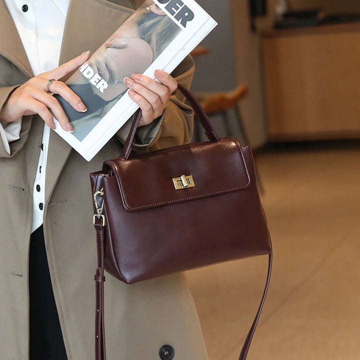 Worn by model, a brown Giselle leather tote bag with a top handle and shoulder strap held by a woman in a trench coat