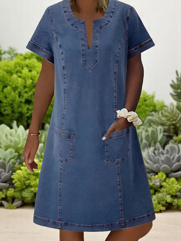 Front view of a woman wearing a soft denim V-neck dress with patch pockets, standing among green succulents