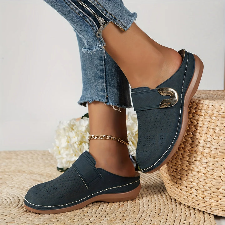 Worn by model, a pair of navy orthopaedic wedge sandals with gold buckle and white stitching on a woven mat