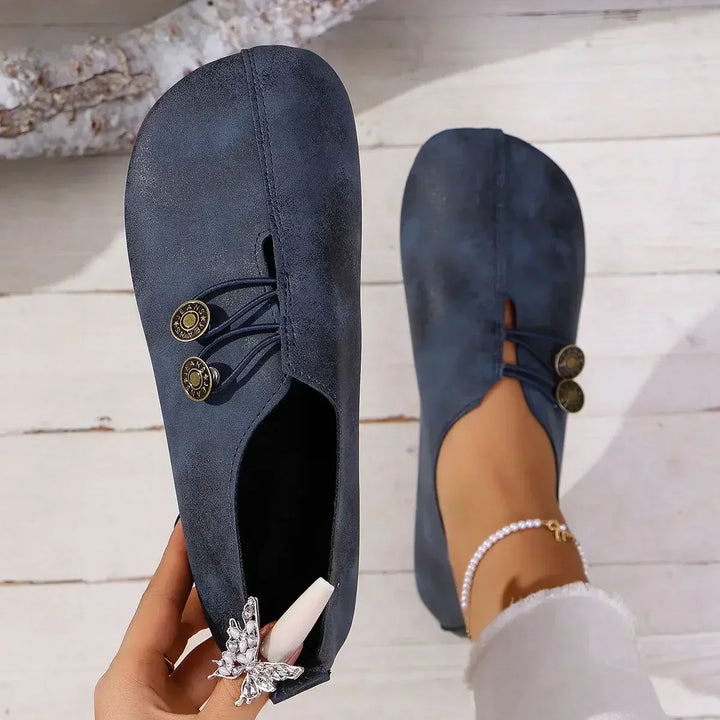Top view of two blue soft suede loafers, one held by hand and one worn by model with a pearl anklet