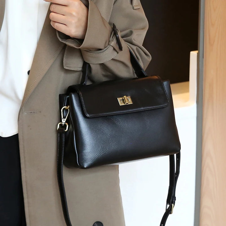 Worn by model, a black Giselle leather tote bag with a top handle and shoulder strap held by a woman in a trench coat