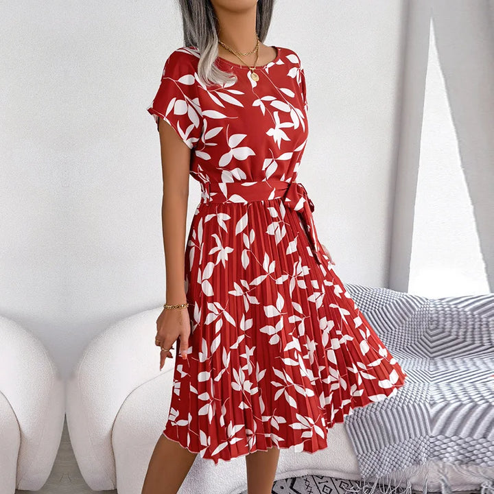 Front view of a model wearing a red floral pleated maxi dress with white leaf print and a tie waist