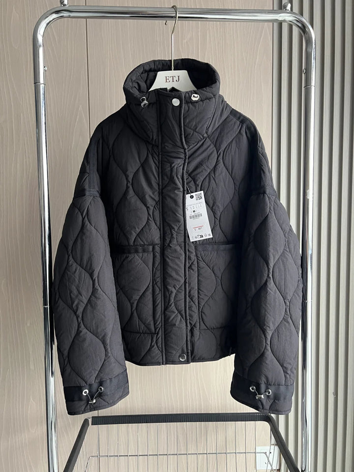 Front view of a black quilted cotton jacket with a high neck, two front pockets and tie waist on a hanger