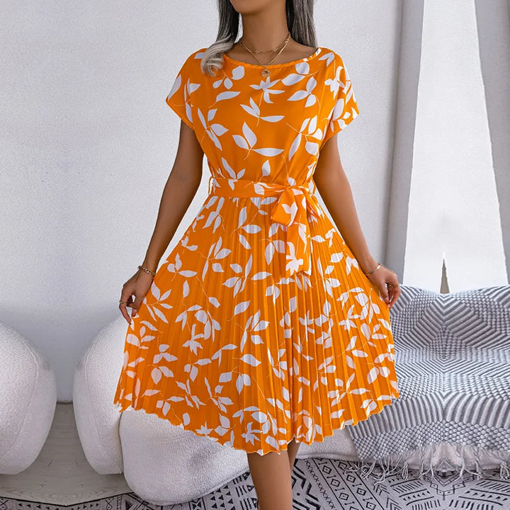 Front view of a model wearing an orange floral pleated maxi dress with white leaf print and a tie waist