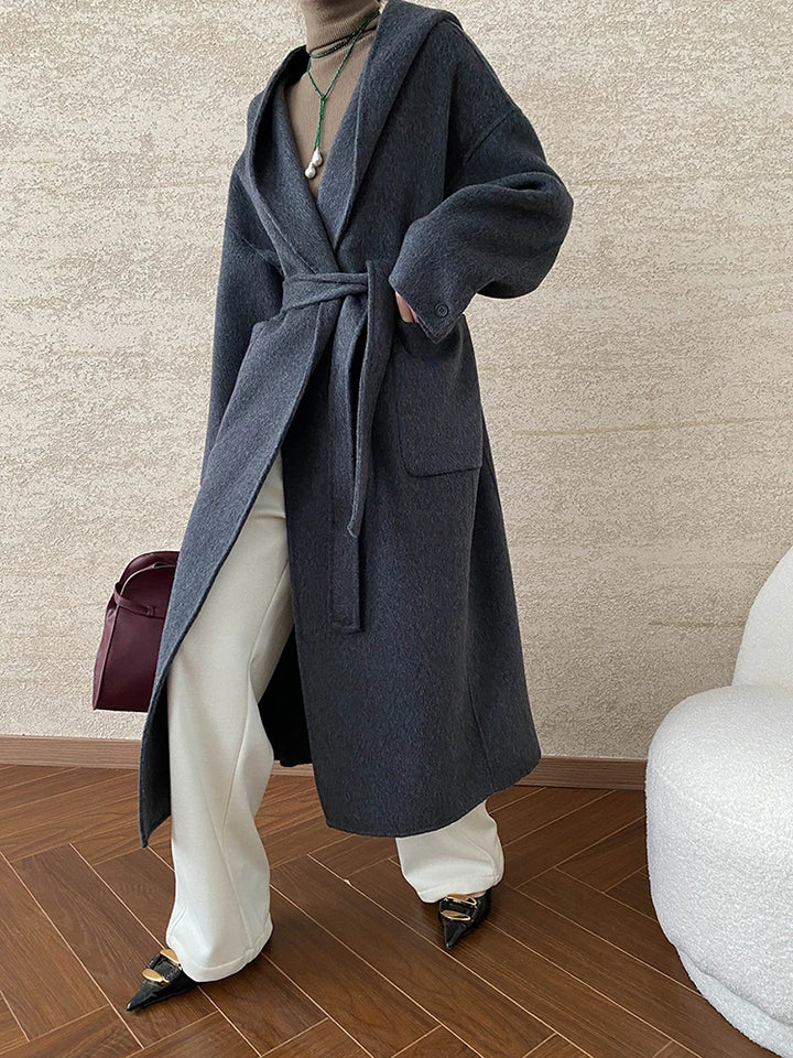 Side view, model wearing a long belted hooded wool blend wrap coat in charcoal with cream trousers