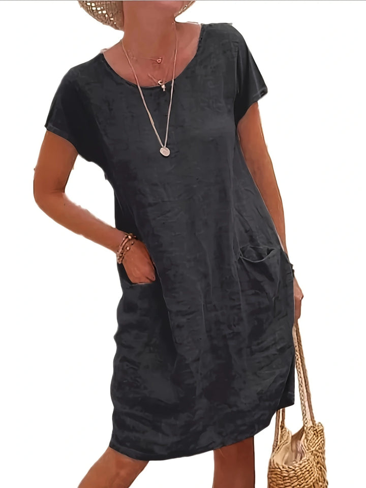 Front view of a blue loose cotton linen dress, worn by model, with hands in pockets and a woven bag