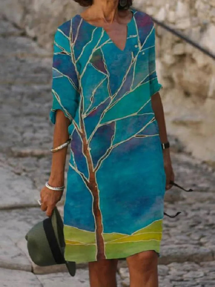 Front view worn by model of a blue vintage printed summer dress with a brown tree and green landscape design