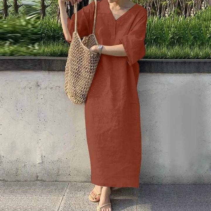 Three quarter view of a model wearing the cotton linen V-neck maxi dress in terracotta, with a net bag and sandals