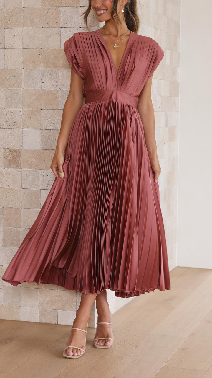 Front view of a model wearing an elegant pleated dress, smiling with hands gently touching the skirt