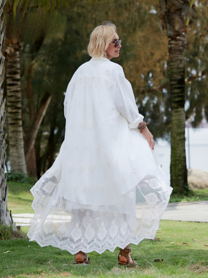 Back view of a blonde model wearing a white lace dress with an elegant overlay walking on grass
