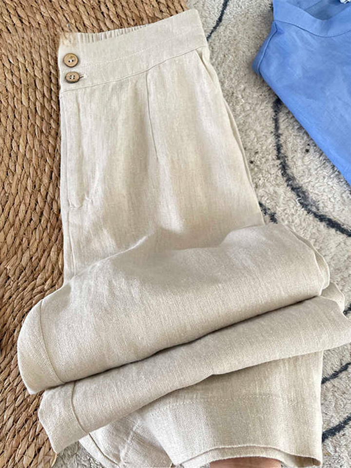Flat lay of beige cotton linen casual pants with button details and folded legs, beside a blue garment
