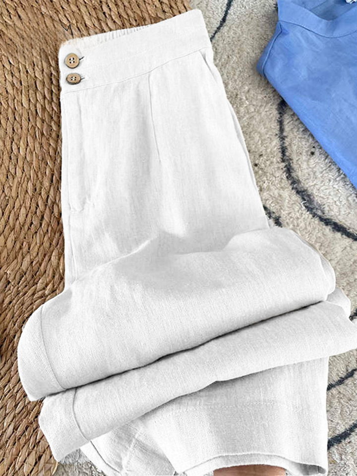 Flat lay of white cotton linen casual pants with button details and folded legs, next to a blue top
