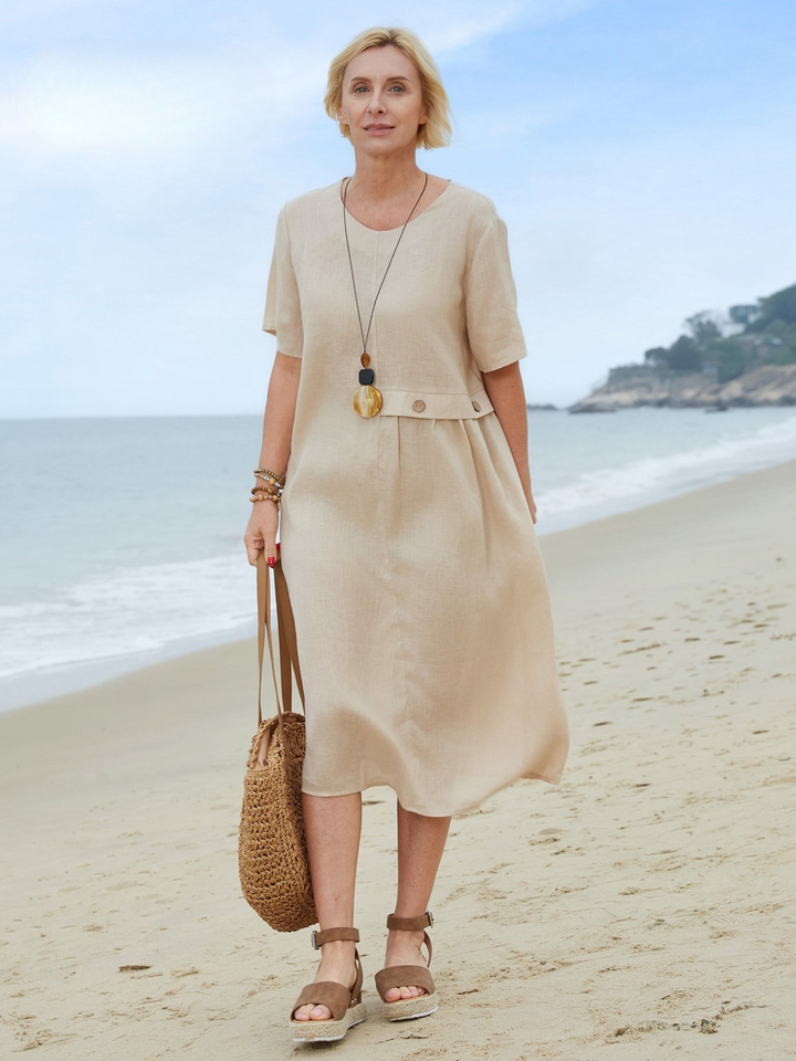 Front view of a blonde model wearing a natural linen pocket dress, holding a woven bag on a sandy beach