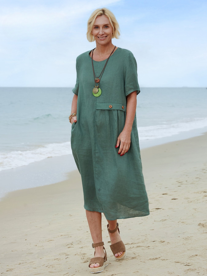 Front view of a blonde model smiling in a natural linen pocket dress, standing on a sandy beach