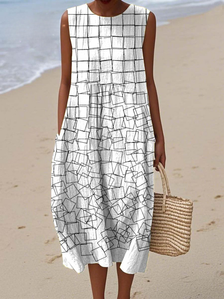 Front view of a model wearing a white geometric pattern wrap dress with black squares, holding a straw bag on a beach