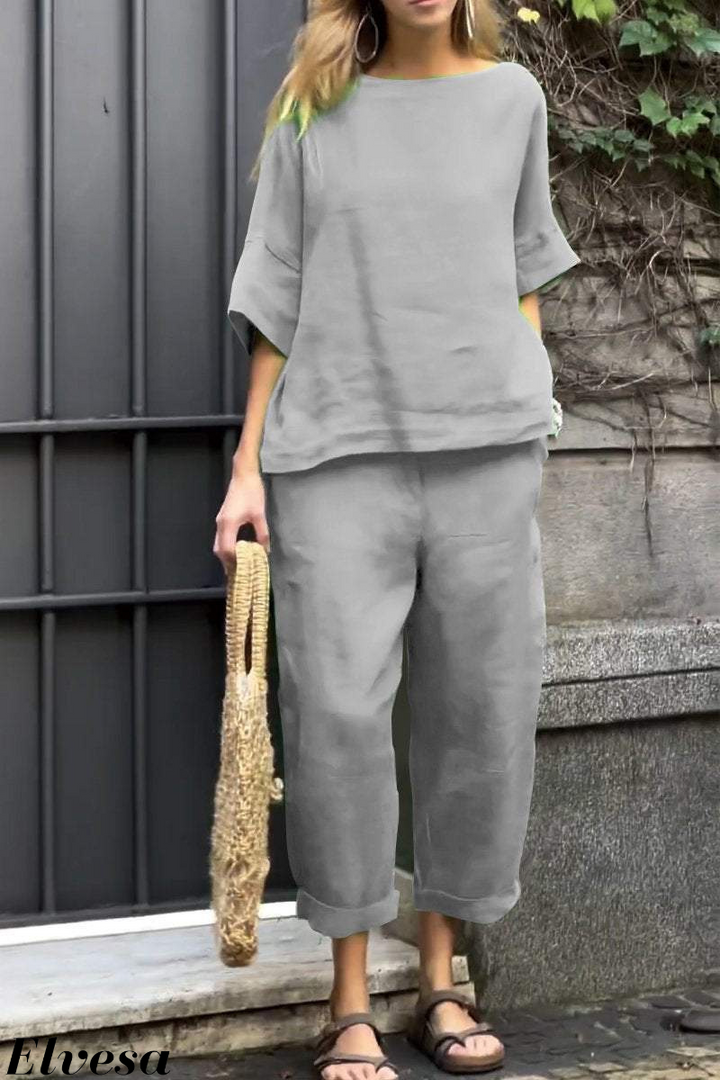 Front view, model wearing a grey two piece set with a boat neck top and cuffed pants, holding a woven bag