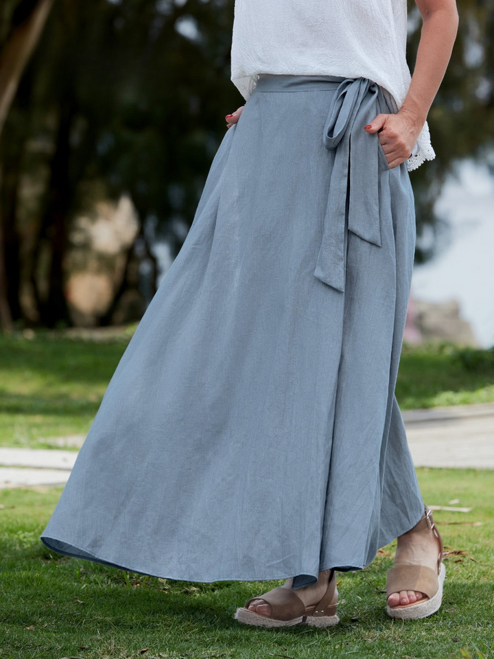 Side view of a model wearing the blue wrap maxi skirt with an adjustable tie and pockets, standing on grass