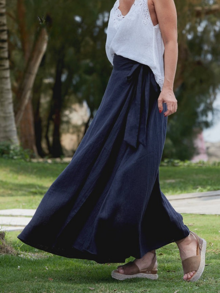 Three quarter view of a model in a blue linen wrap maxi skirt with adjustable tie, walking on grass