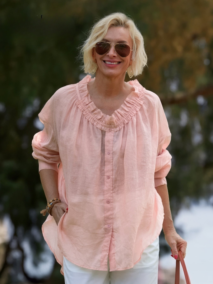 Front view of a blonde model wearing a loose fitting pink linen shirt blouse with a ruffled neckline