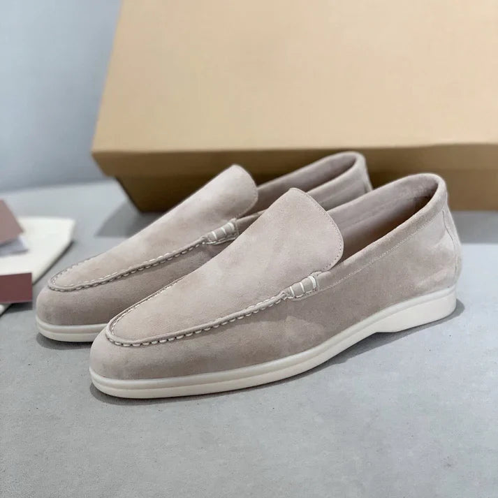 Three quarter view of two light beige suede loafers with white soles on a grey surface and a shoe box behind