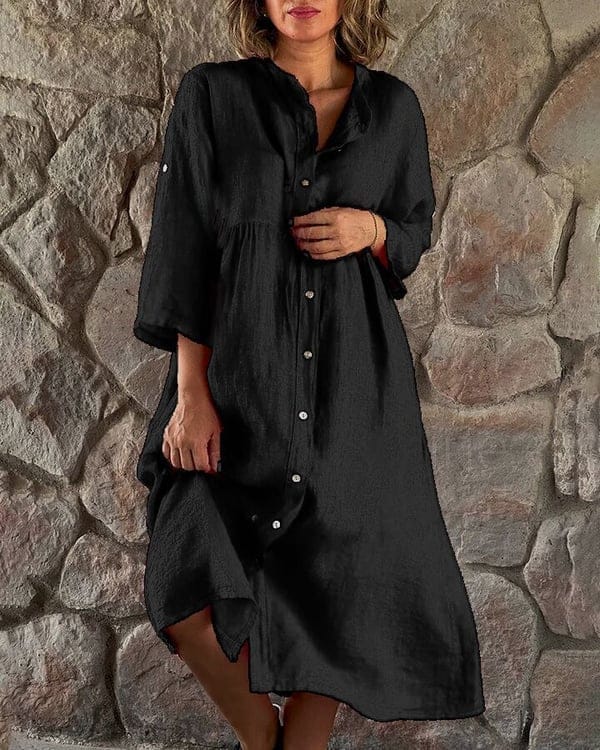 Front view of a model wearing a black loose shirt midi dress with white buttons against a stone wall