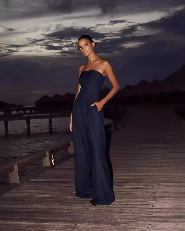 Front view of a model wearing a strapless navy Luxe Flow Jumpsuit on a wooden pier at dusk