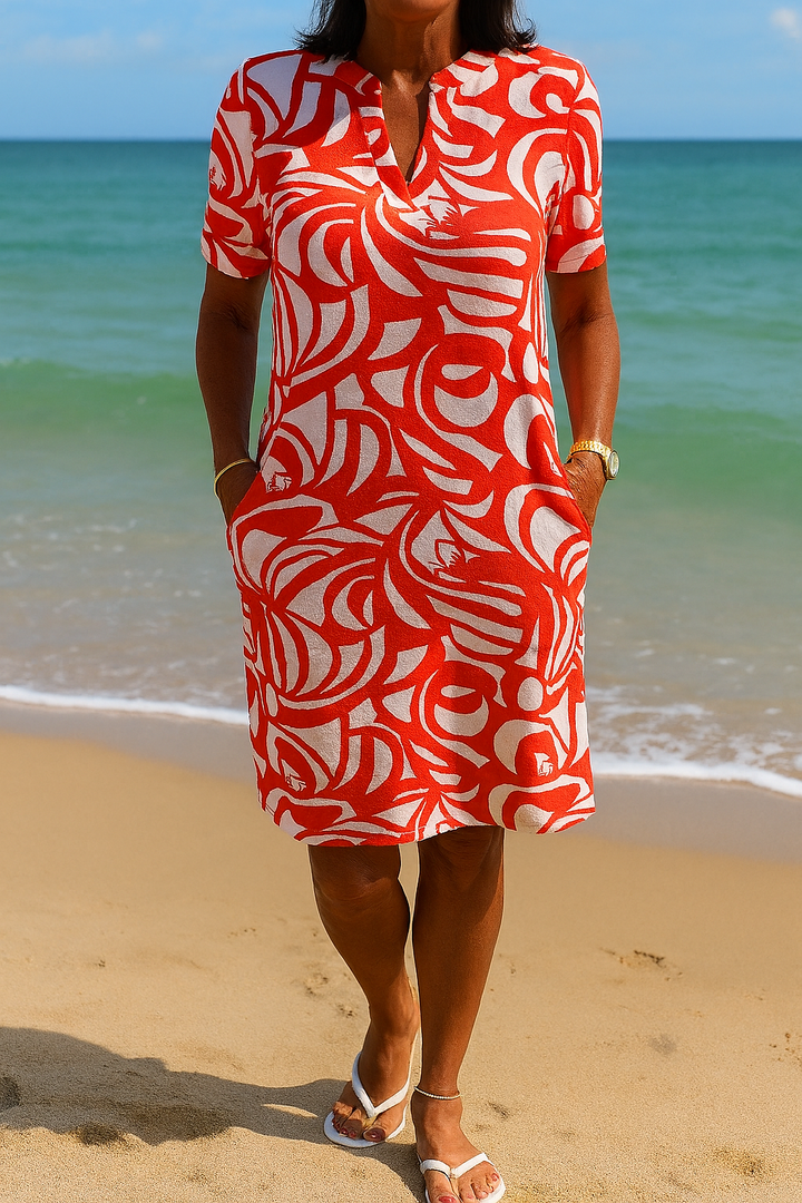 Front view of a model wearing a red and white printed V-neck summer shirt dress with hands in pockets on a beach