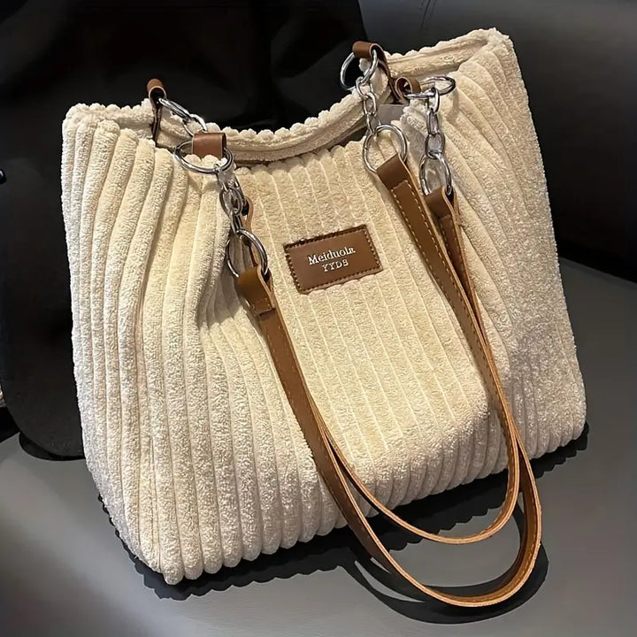 Front view of a cream corduroy tote bag with brown straps and silver chain details on a dark chair