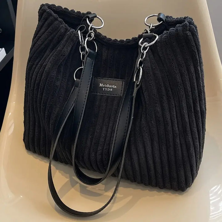 Front view of a black corduroy tote bag with black straps and silver chain details on a light chair