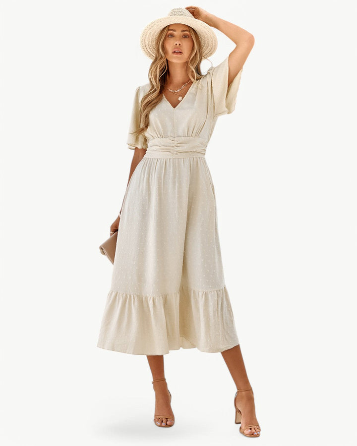 Front view, worn by model, a beige V-neck maxi dress with short sleeves, shirred waist and ruffle hem, holding a clutch