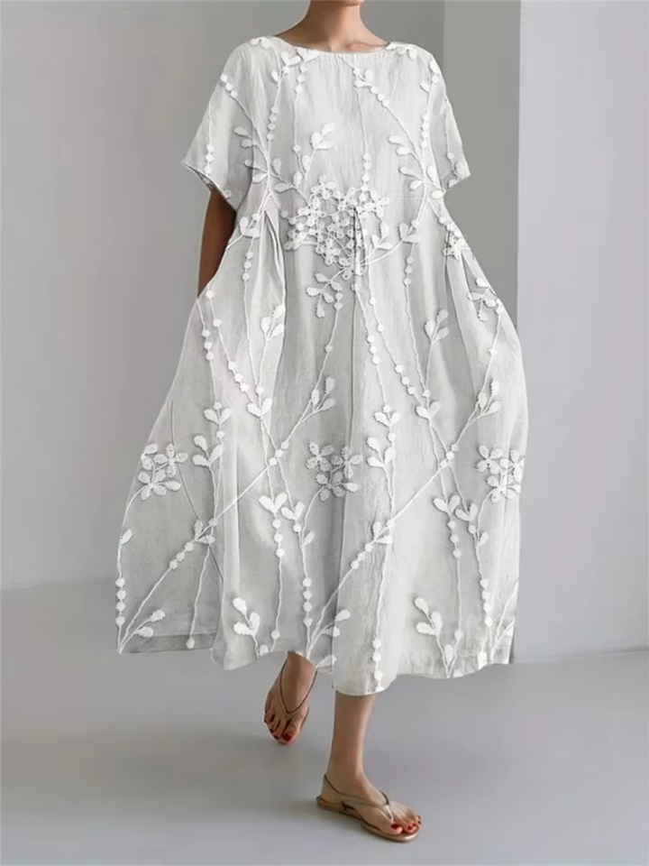 Elegant long dress worn by model, three quarter view, showing white vine embroidery on light beige linen blend fabric