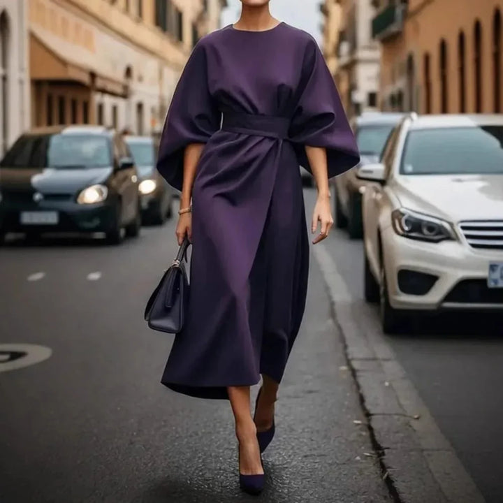 Front view of a model in an elegant belted kimono sleeve midi dress with a black handbag on a street, walking