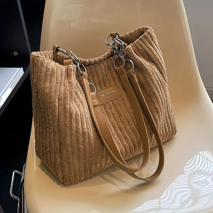 Three quarter view of a tan corduroy tote bag with light brown straps and silver chains on a cream chair