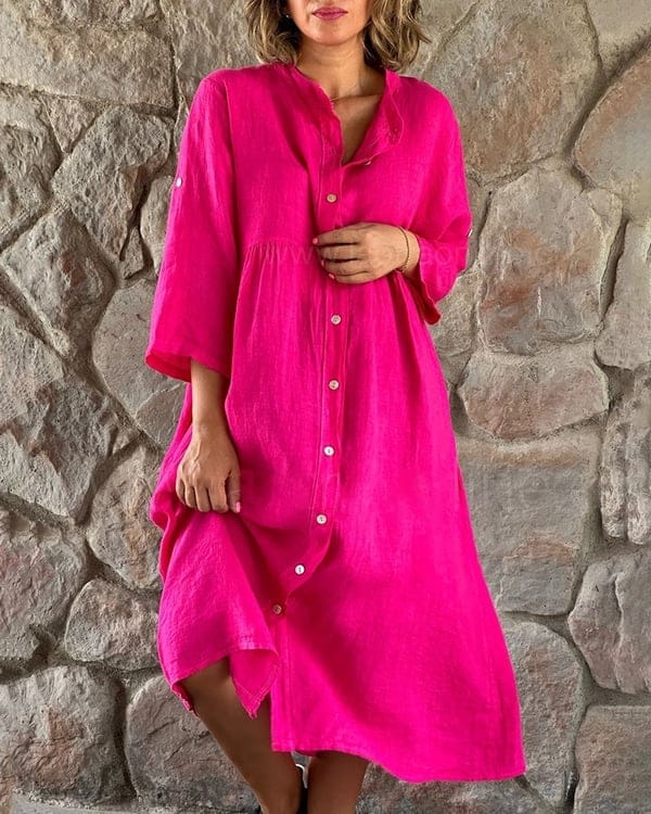 Front view of a model wearing a bright pink loose shirt midi dress with white buttons against a stone wall