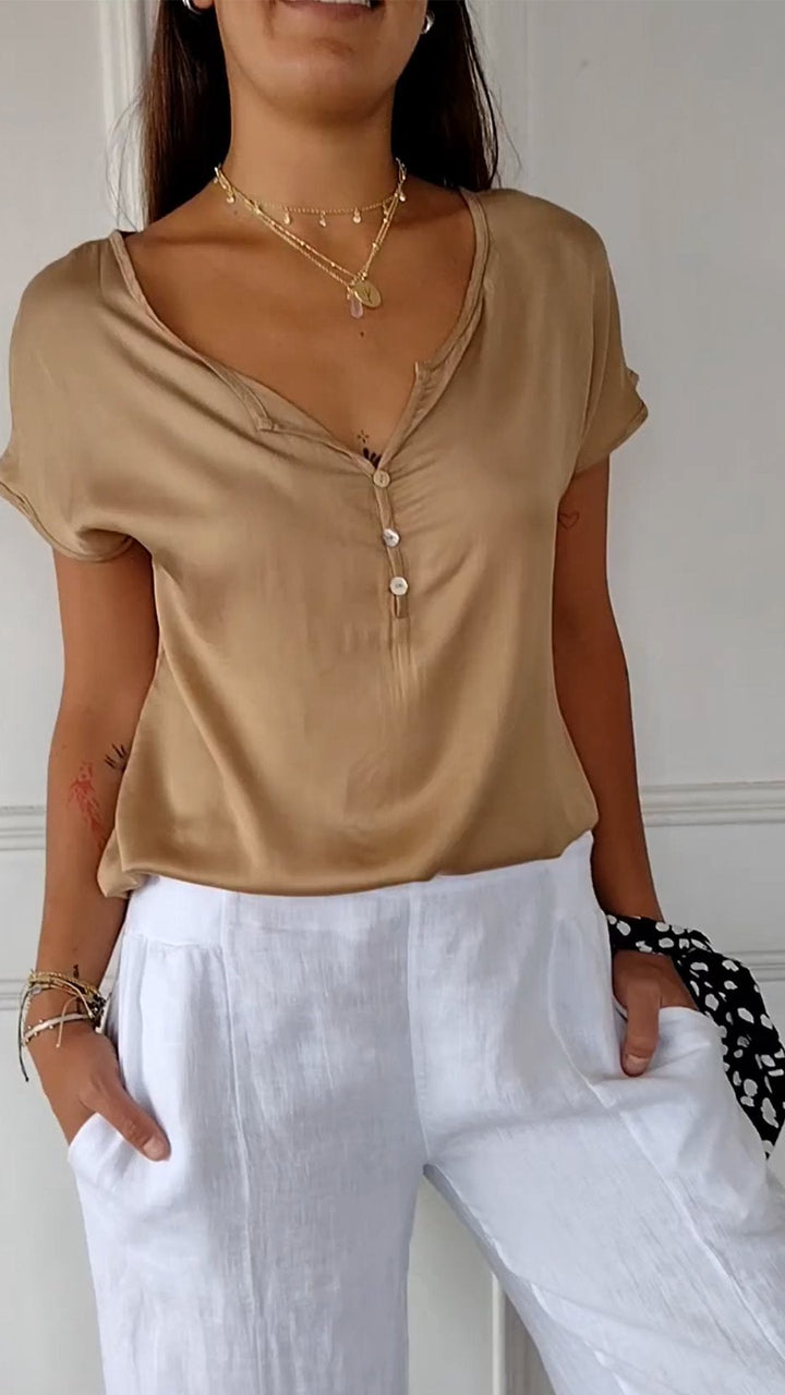 Front view of a woman wearing a tan V-neck tee with white buttons and white linen trousers, hands in pockets