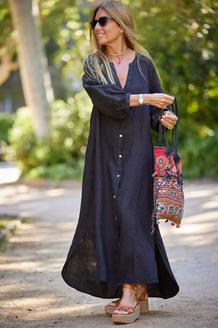 Front view, model smiling in a dark retro loose maxi dress with long sleeves and a colourful embroidered handbag