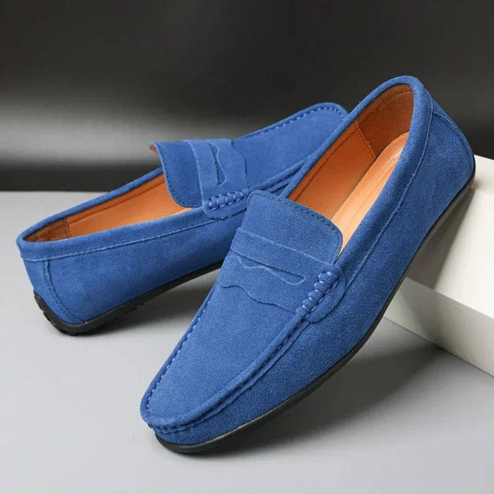 Pair of blue suede loafers, three quarter view, with a black sole and tan interior