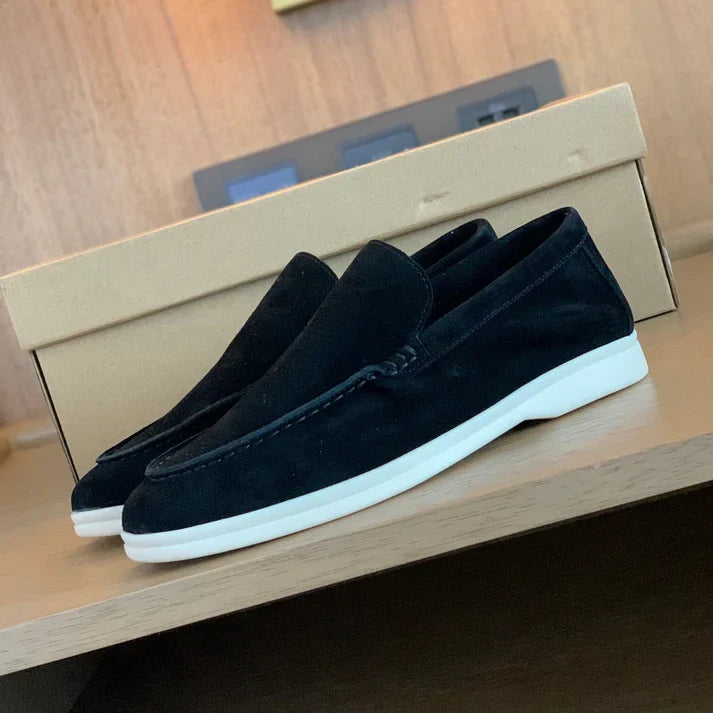 Front view of black suede loafers with white soles on a wooden surface, a shoe box in the background