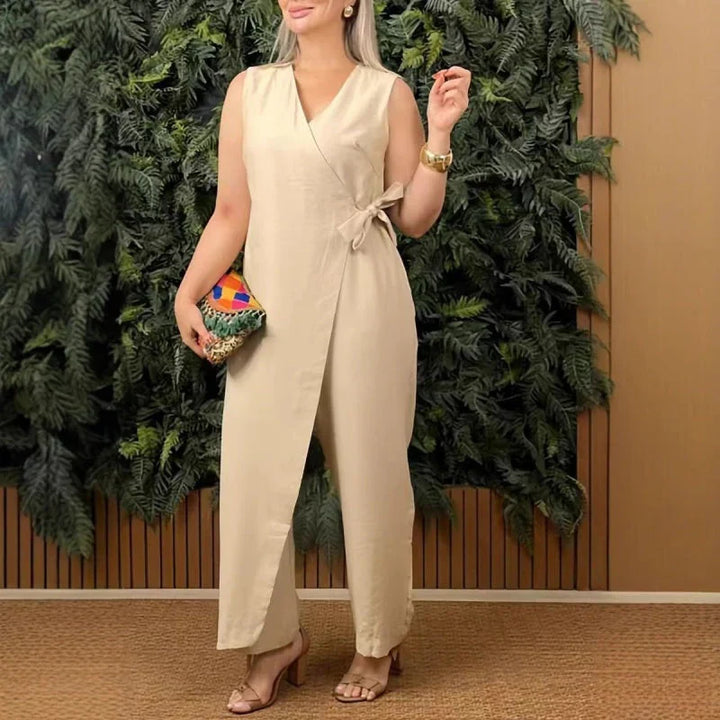 Front view of a model wearing a stone V-neck jumpsuit with a side tie, holding a colourful woven clutch